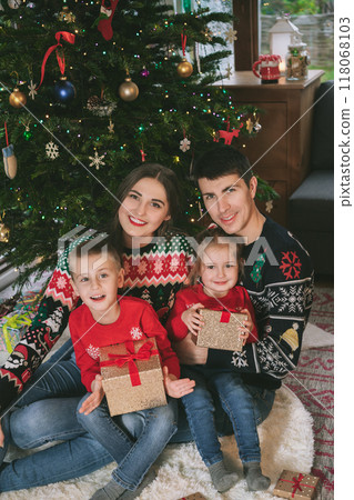 Smiling family portrait in festive sweaters sitting with gifts under Christmas tree at home during holiday. Happy parents and kids cuddling, feeling excited. Family time together. Happy childhood. Smiling family portrait in festive sweaters sitting with gifts under Christmas tree at home during holiday. Happy parents and kids cuddling, feeling excited. Family time together. Happy childhood. 118068103