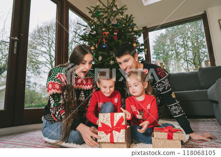 Happy family in festive sweaters sitting under Christmas tree at home. Emotional parents and kids feeling excited unwrapping Christmas gifts, having fun, Family time, childhood miracle time concept. Happy family in festive sweaters sitting under Christmas tree at home. Emotional parents and kids feeling excited unwrapping Christmas gifts, having fun, Family time, childhood miracle time concept. 118068105