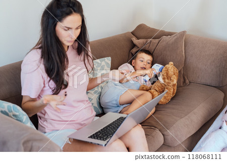 Focus on sick boy measuring temperature of his toy bear with non-contact digital infrared thermometer while his mother has online video chat with pediatrician doctor on pc. Telemedicine, telehealth. Focus on sick boy measuring temperature of his toy bear with non-contact digital infrared thermometer while his mother has online video chat with pediatrician doctor on pc. Telemedicine, telehealth. 118068111