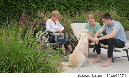 Family members, including a senior woman in a wheelchair, enjoy a relaxed moment with their Golden Retriever outdoors in a serene garden. 118069574