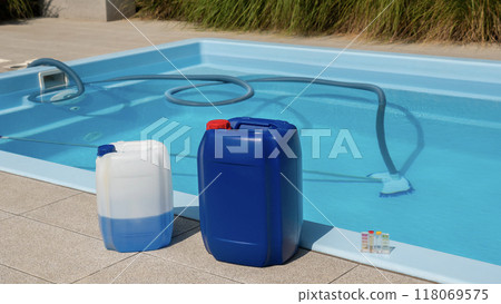 A poolside scene featuring chemical containers, a submerged cleaning hose in the pool, and a water testing kit, highlighting pool maintenance routines. 118069575
