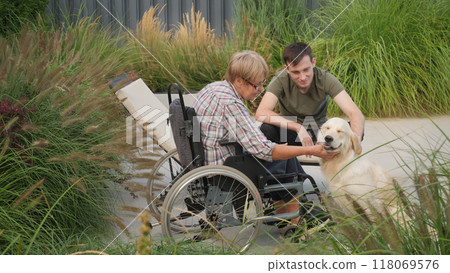 A senior woman in a wheelchair and a young man pet their Golden Retriever outdoors, sharing a joyful moment in a lush garden. 118069576