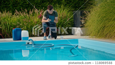 Pool technician using a tablet by the pool, surrounded by chemical containers and greenery, focused on water quality. 118069588