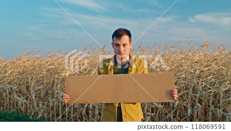 A man holding a blank cardboard sign stands in a dry cornfield, wearing a yellow shirt against a blue sky. A man holding a blank cardboard sign stands in a dry cornfield, wearing a yellow shirt against a blue sky. 118069591