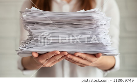 A person grips a massive pile of papers, showcasing the challenges of managing paperwork in an office environment. The bright light emphasizes the seriousness of the task 118069749