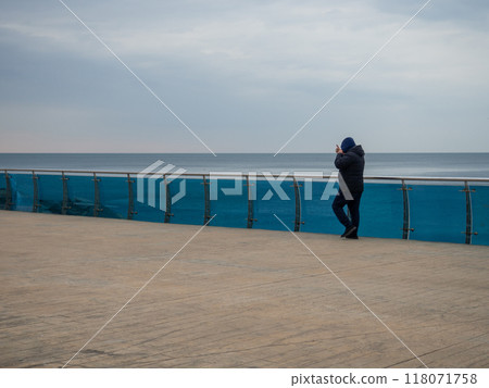 A man walks along the bridge along the seashore. Walking in the resort in the winter season.  man in the hood speaks on the phone. Coast of the Black Sea. 118071758