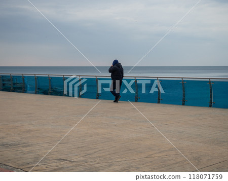 A man walks along the bridge along the seashore. Walking in the resort in the winter season. The man in the hood speaks on the phone. Coast of the Black Sea. 118071759