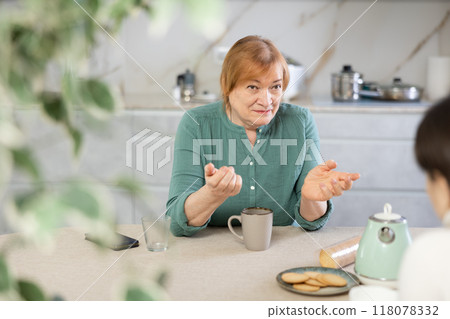 Annoyed elderly woman with cup of tea in her hands in kitchen listening to her friend Annoyed elderly woman with cup of tea in her hands in kitchen listening to her friend 118078332