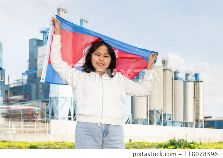 Happy young woman with flag of Cambodia against background of factory 118078396