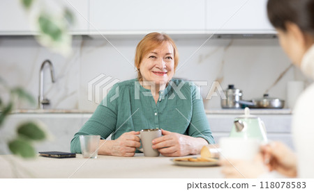 In kitchen at table, elderly smiling woman communicates with friend and drinks coffee or tea 118078583