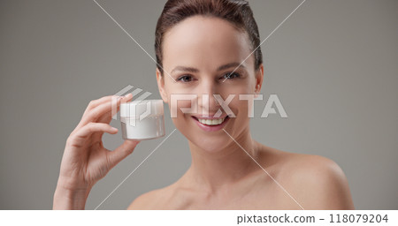 Beauty, health, cosmetics, anti-aging therapy and skin care concept - young beautiful brunette Caucasian woman holding a jar of moisturizer in her hands and looking at the camera 118079204