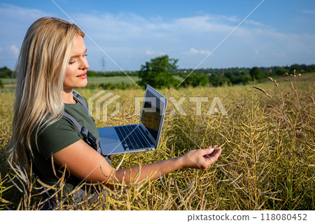 A woman farmer checking the quality of rapeseed, standing at the rapeseed field and holding a laptop A woman farmer checking the quality of rapeseed, standing at the rapeseed field and holding a laptop 118080452