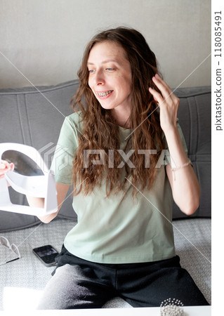 beautiful young woman with long wavy hair doing her hair and admiring her reflection while sitting on the sofa at home beautiful young woman with long wavy hair doing her hair and admiring her reflection while sitting on the sofa at home 118085491