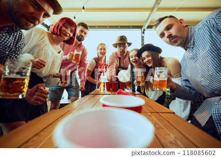 Group of people cheerfully playing beer pong, having fun and bar while celebrating beer festival. Bavarian traditions 118085602