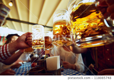 Focus on lager beer mugs clinking, with blurred people on background, Group of people meeting at pub for cheerful celebration 118085748