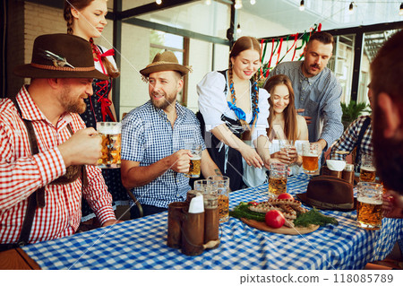 Oktoberfest joy. Men and women, friends sitting at table at local bar, enjoying beer. Celebration with Bavarian traditions 118085789