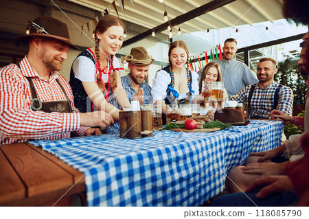 Two women, waitress dressed in Bavarian clothes serving beer to a group of friends meeting at pub for cheerful celebration Two women, waitress dressed in Bavarian clothes serving beer to a group of friends meeting at pub for cheerful celebration 118085790