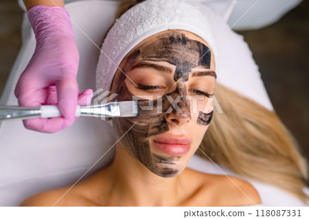cosmetologist applying black mask on pretty woman face wearing black gloves, gorgeous woman in spa having facial procedures cosmetologist applying black mask on pretty woman face wearing black gloves, gorgeous woman in spa having facial procedures 118087331