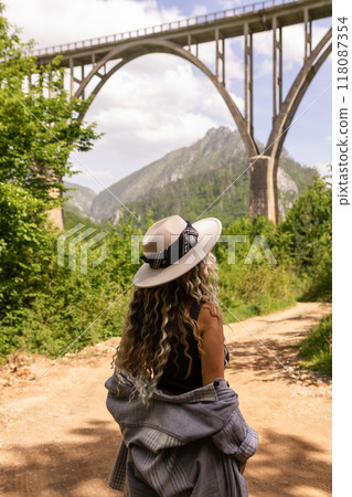 A girl in a hat stands against the backdrop of a beautiful arched bridge over the Tara River, a popular tourist attraction in Montenegro. A girl in a hat stands against the backdrop of a beautiful arched bridge over the Tara River, a popular tourist attraction in Montenegro. 118087354