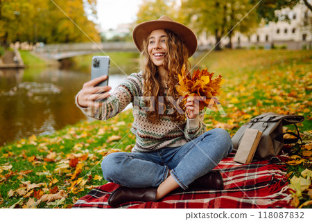 Selfie time. Beautiful woman takes selfie on smartphone in hat on meadow in autumn park on plaid. Selfie time. Beautiful woman takes selfie on smartphone in hat on meadow in autumn park on plaid. 118087832