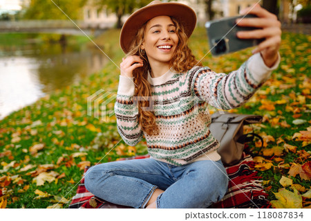 Selfie time. Beautiful woman takes selfie on smartphone in hat on meadow in autumn park on plaid. Selfie time. Beautiful woman takes selfie on smartphone in hat on meadow in autumn park on plaid. 118087834