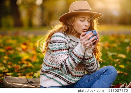 Curly woman with thermos in clearing among fallen leaves in autumn park, enjoying hot drink. Curly woman with thermos in clearing among fallen leaves in autumn park, enjoying hot drink. 118087870