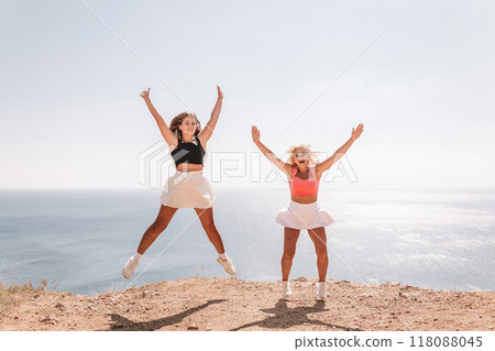 Two women are jumping in the air on a beach. The sky is blue and the ocean is in the background. Scene is joyful and energetic. Two women are jumping in the air on a beach. The sky is blue and the ocean is in the background. Scene is joyful and energetic. 118088045