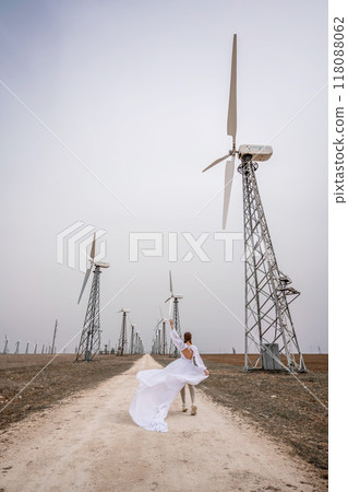 A woman in a white dress is walking down a dirt road in front of a row of wind turbines. 118088062