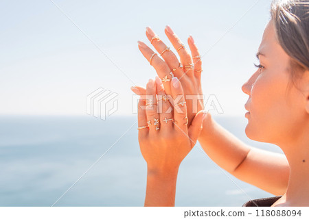 A woman is wearing a lot of rings on her hands and is looking out at the ocean. The rings are of various sizes and designs, and the woman's hands are spread wide apart. 118088094