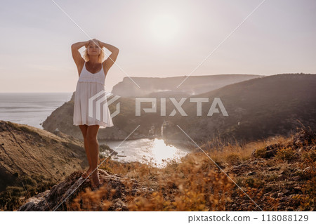 A blonde woman stands on a hill overlooking the ocean. She is wearing a white dress and she is enjoying the view. 118088129