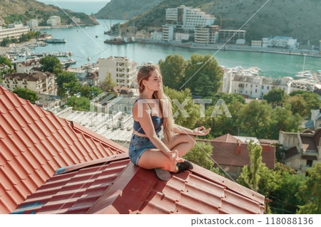 Woman sits on rooftop, enjoys town view and sea mountains. Peaceful rooftop relaxation. Below her, there is a town with several boats visible in the water. Rooftop vantage point. 118088136