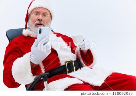 Santa Claus, looking surprised, stares at his smartphone while holding cup in his other hand against white studio background. 118092083