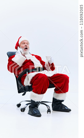 Santa Claus is sitting in office chair, talking on phone with surprised expression while holding cup against white studio background. Santa Claus is sitting in office chair, talking on phone with surprised expression while holding cup against white studio background. 118092085