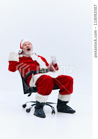 Santa Claus sitting on rolling office chair, holding cup in one hand, and talking one phone with excited expression against white background. Santa Claus sitting on rolling office chair, holding cup in one hand, and talking one phone with excited expression against white background. 118092087