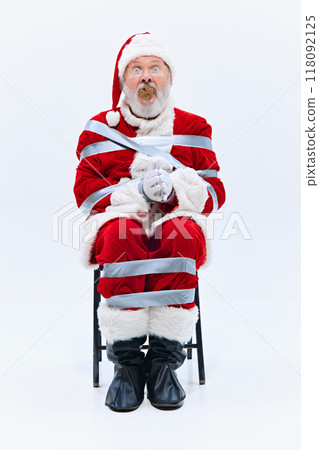 Santa is wrapped tightly in duct tape with pinecone stuffed in his mouth, his eyes wide in disbelief as he sits immobilized on chair. Santa is wrapped tightly in duct tape with pinecone stuffed in his mouth, his eyes wide in disbelief as he sits immobilized on chair. 118092125