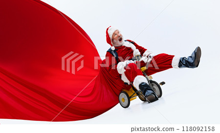 Joyful, shocked Santa Claus riding tricycle with a big red bag of gifts flowing behind against white studio background. 118092158
