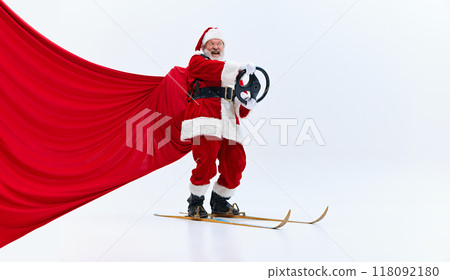 Santa Claus skiing with arms raised, holding ski poles, red cape billowing behind him, smiling broadly against white studio background. 118092180