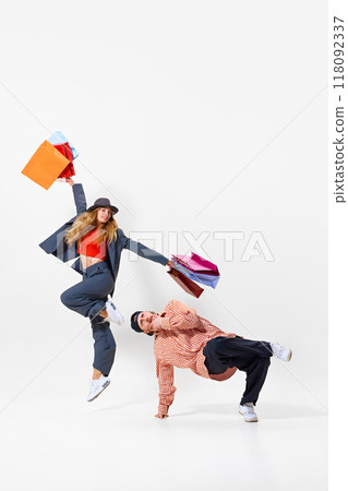 Happy young woman in a suit jumping with multicolored shopping bags, while man performing breakdance pose. Shopping humorous activity 118092337