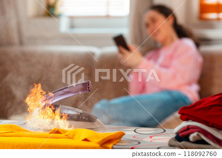 Firing Iron is on ironing board. In the background, a woman is sitting resting on the couch and using a smartphone. The concept of household fire danger 118092760