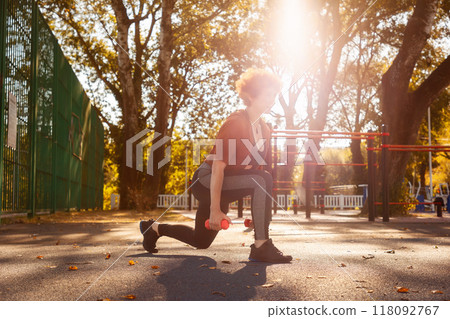 Adult Caucasian woman wearing sport clothes squats holds dumbbells Sunlight in background. Concept of outdoor training and healthy lifestyle Adult Caucasian woman wearing sport clothes squats holds dumbbells Sunlight in background. Concept of outdoor training and healthy lifestyle 118092767