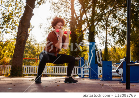 Adult Caucasian woman with makeup squatting hardly holds dumbbells in sports park. Concept of healthy lifestyle and fitness Adult Caucasian woman with makeup squatting hardly holds dumbbells in sports park. Concept of healthy lifestyle and fitness 118092769