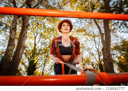 Bottom view through horizontal bar of mid adult Caucasian woman training with sports elastic band. Tree crowns in background. Concept of sport Bottom view through horizontal bar of mid adult Caucasian woman training with sports elastic band. Tree crowns in background. Concept of sport 118092777