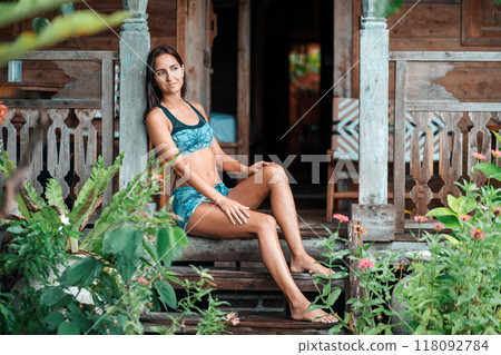 A beautiful tanned woman in summer clothes poses sitting on the steps of a wooden house, surrounded by tropical plants. Summer vacation and travel concept 118092784