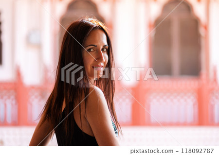 A beautiful woman poses smiling. In the background, pink arches and Windows, Oriental architecture. Rear view. Travel concept 118092787