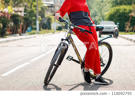 Sports lifestyle. A woman in red sportswear, posing leaning on a Bicycle. In the background, an empty road and street. Eco-friendly transport concept. Close up of legs Sports lifestyle. A woman in red sportswear, posing leaning on a Bicycle. In the background, an empty road and street. Eco-friendly transport concept. Close up of legs 118092795