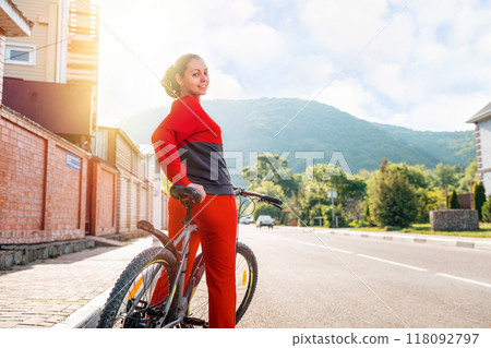 Sports lifestyle. Beautiful smiling young woman in sportswear posing sitting on a Bicycle. Rear view. In the background, an empty street. Sunlight. Copy space Sports lifestyle. Beautiful smiling young woman in sportswear posing sitting on a Bicycle. Rear view. In the background, an empty street. Sunlight. Copy space 118092797