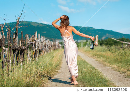 Woman in satin dress goes barefoot in the road, holding the hands of shoes and a bottle of alocohol. Countryside and freedom. Rear view 118092808