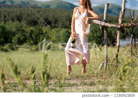 Woman in satin dress standing barefoot on the grass, leaning against the old wooden fence, holding the hands of shoes and a bottle of alocohol. Countryside and freedom Woman in satin dress standing barefoot on the grass, leaning against the old wooden fence, holding the hands of shoes and a bottle of alocohol. Countryside and freedom 118092809