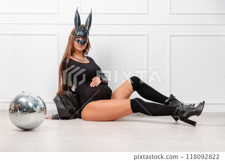 A pregnant woman in a rabbit mask, with long dark hair, in a black bodysuit poses with a mirror ball, holding her stomach with her hands. Studio fashion photography A pregnant woman in a rabbit mask, with long dark hair, in a black bodysuit poses with a mirror ball, holding her stomach with her hands. Studio fashion photography 118092822