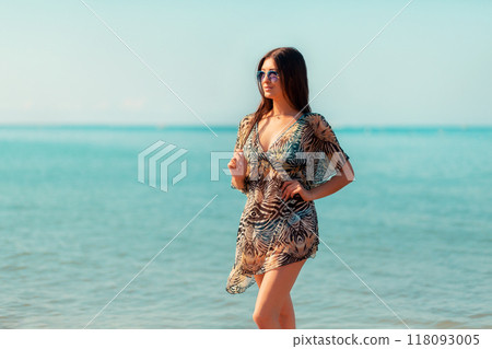 A beautiful woman in a pareo and sunglasses poses against the sea and sky. Concept of beach holidays and summer vacations A beautiful woman in a pareo and sunglasses poses against the sea and sky. Concept of beach holidays and summer vacations 118093005